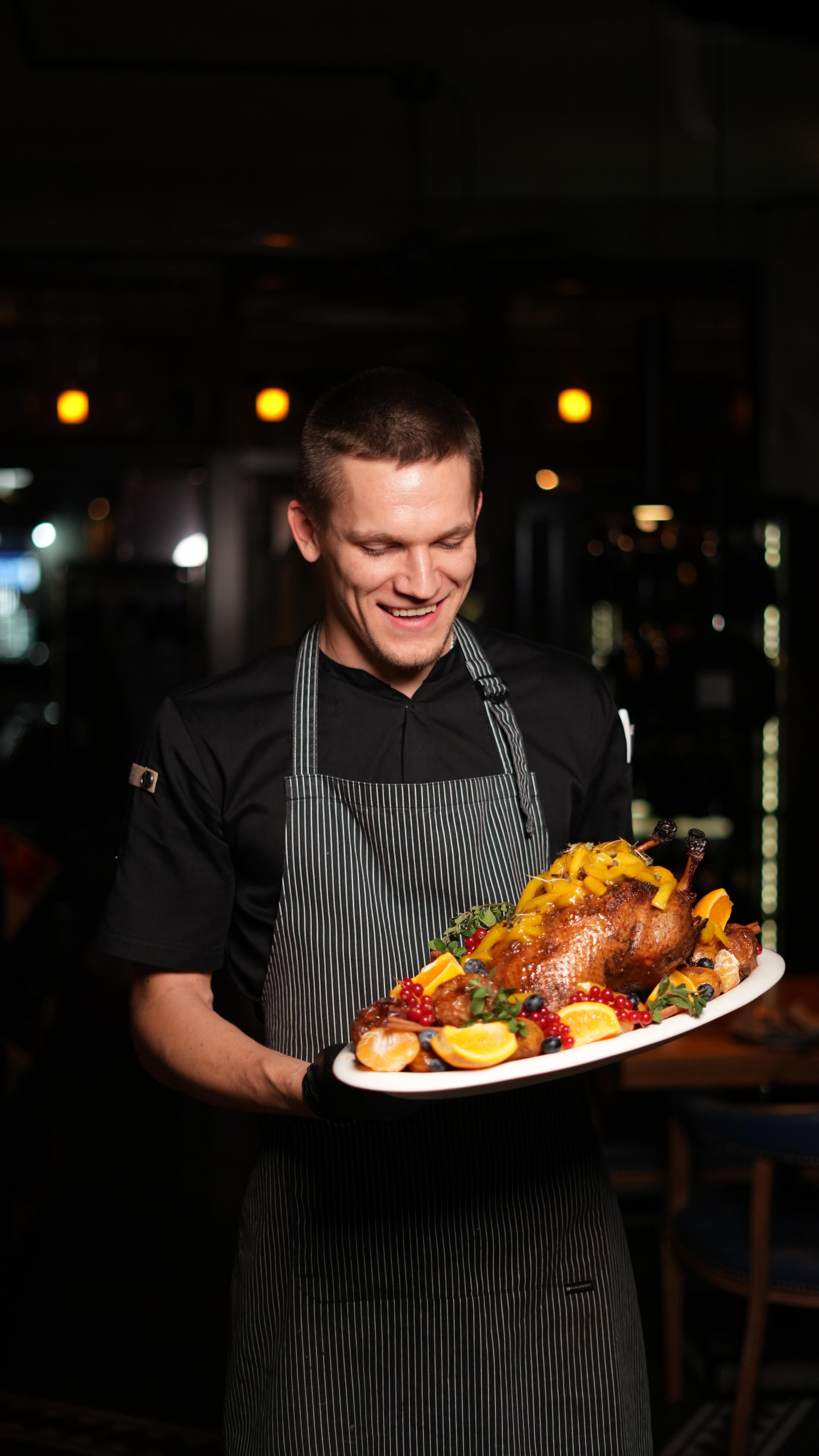 Ein lächelnder Koch mit kurzem Haar hält einen festlich dekorierten Braten auf einem Teller
