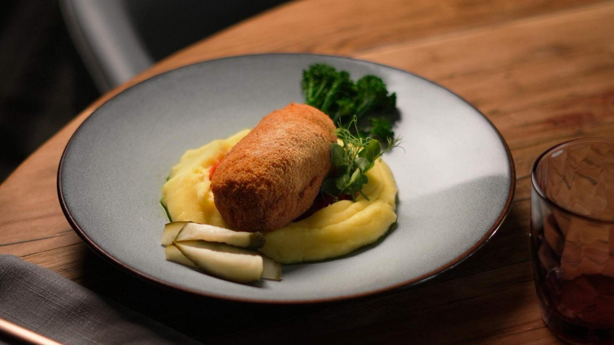 Ein hellbraunes paniertes Röllchen liegt auf cremigem Kartoffelpüree mit Gemüse und frischem Grün